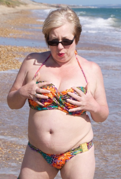 Mature UK woman Speedy Bee wears sunglasses while getting naked at the beach